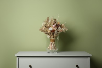 Beautiful dried flower bouquet in glass vase on white chest of drawers near olive wall