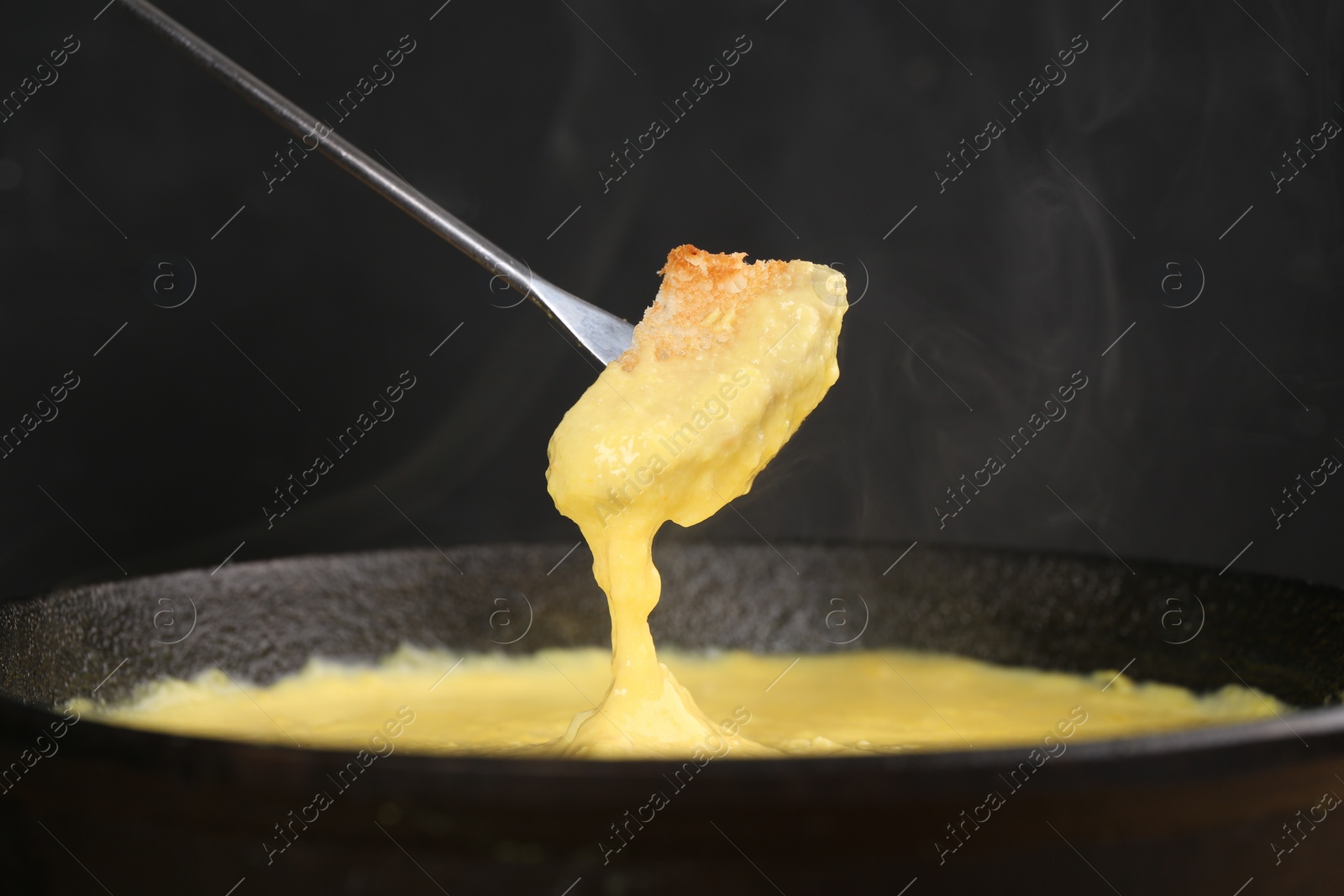 Photo of Dipping piece of bread into fondue pot with tasty melted cheese against dark gray background, closeup