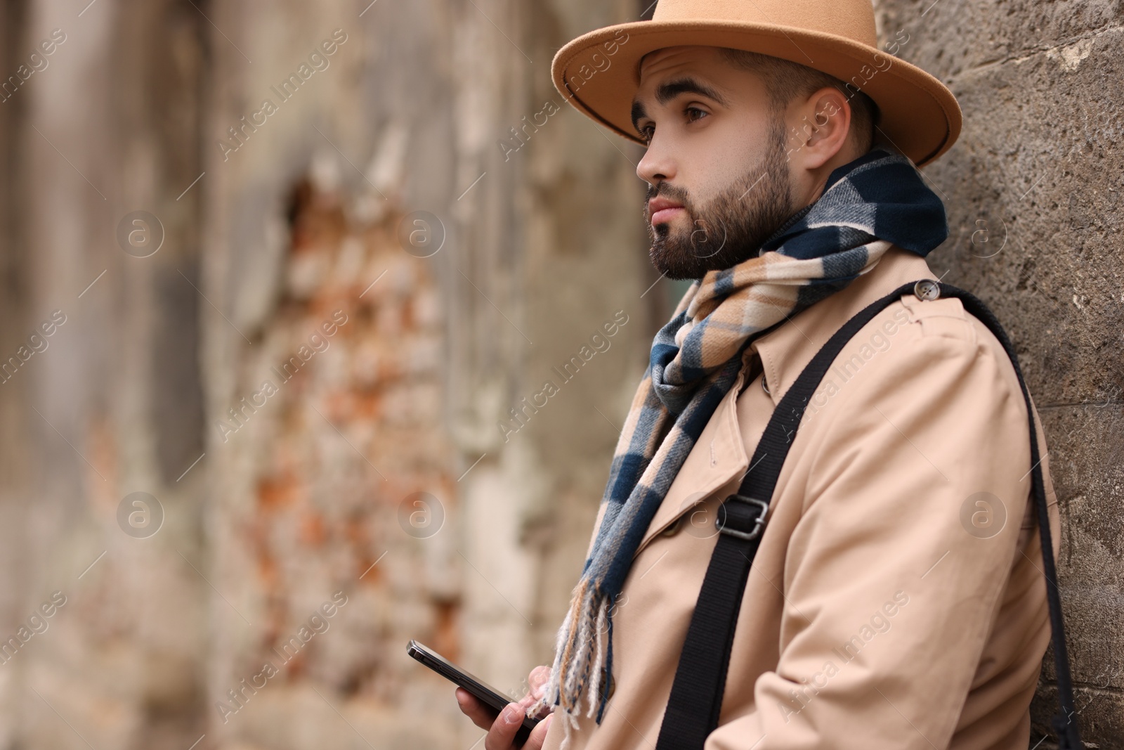 Photo of Handsome man in warm scarf with smartphone near wall outdoors. Space for text