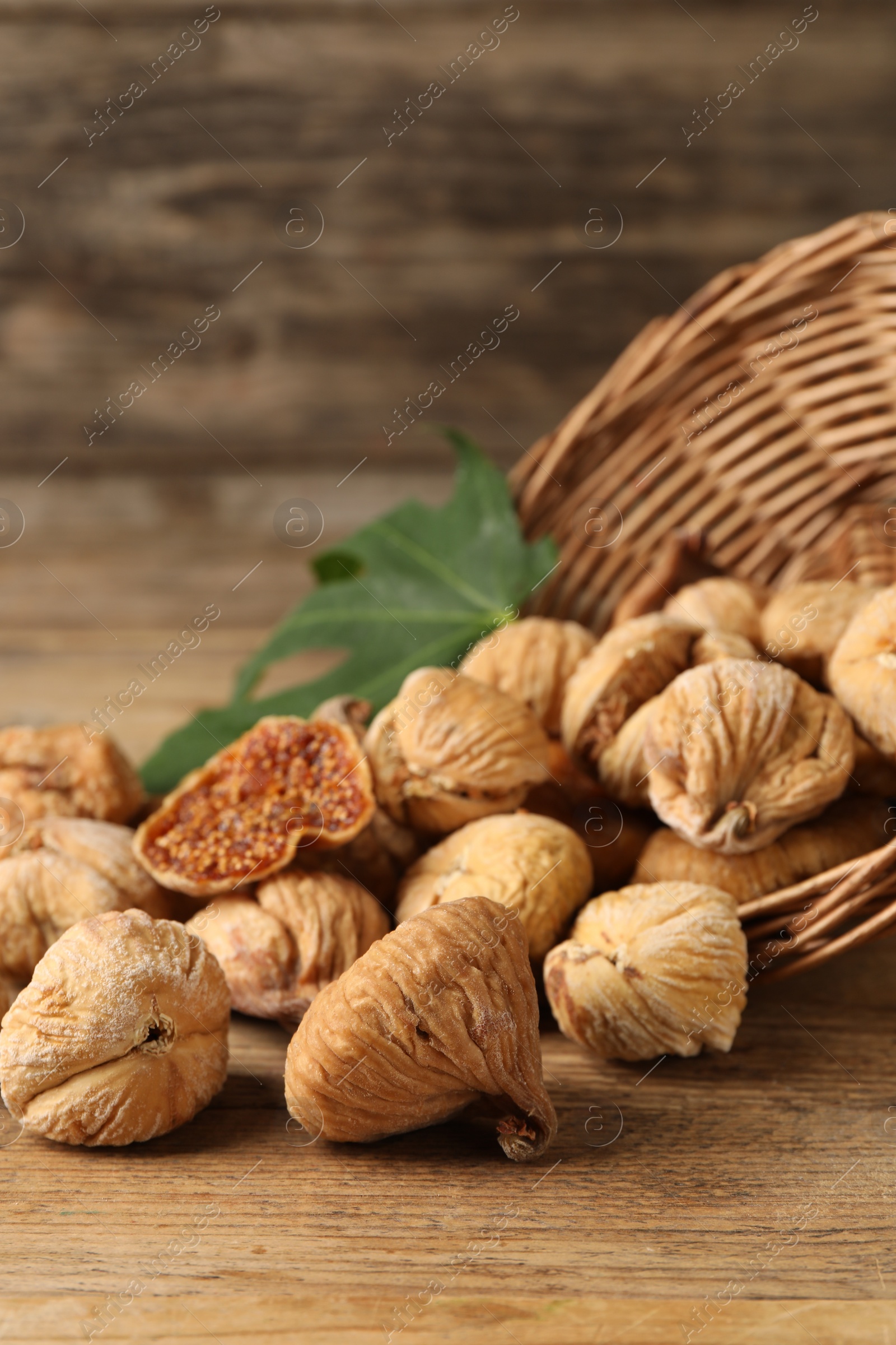 Photo of Overturned wicker basket, dried figs and green leaf on wooden table, closeup