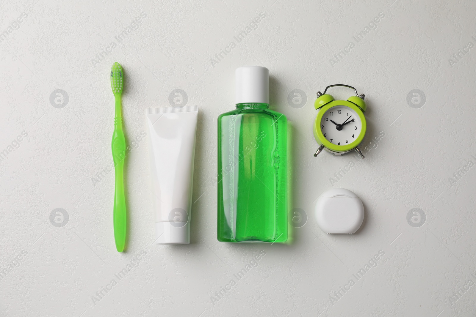 Photo of Fresh mouthwash in bottle, toothbrush, toothpaste, dental floss and alarm clock on light background, flat lay