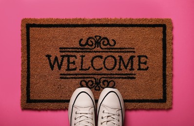 Doormat with word Welcome and stylish sneakers on pink background, flat lay