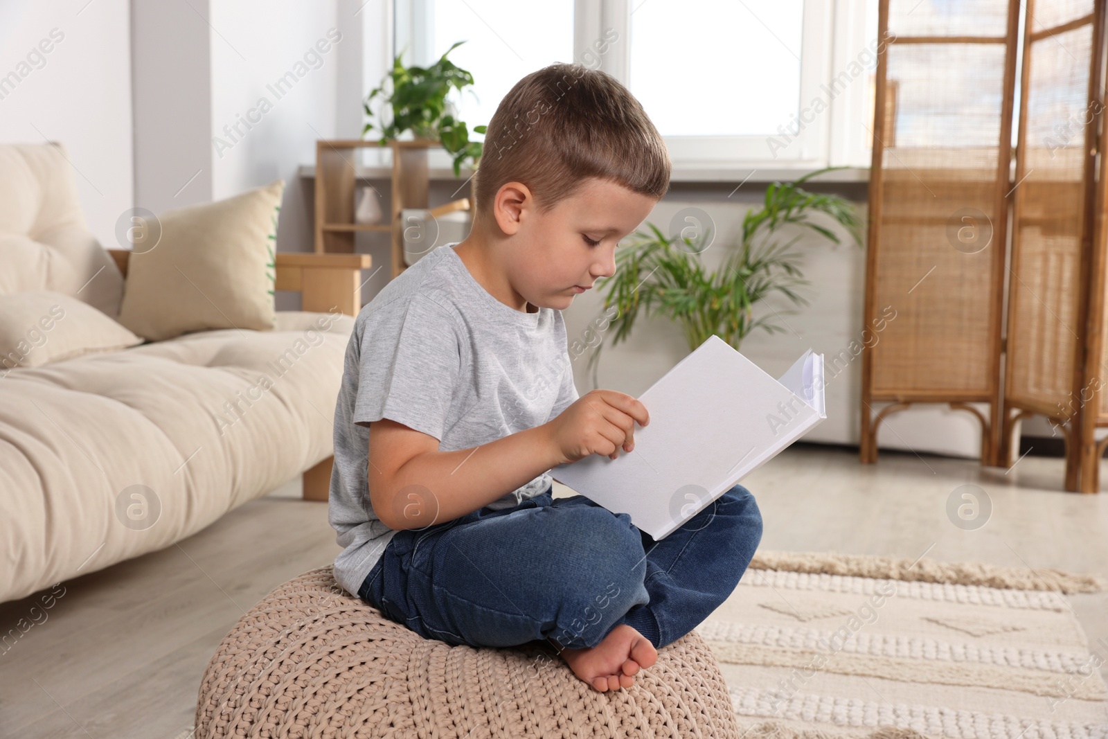 Photo of Boy with poor posture reading book on beige pouf in living room. Symptom of scoliosis