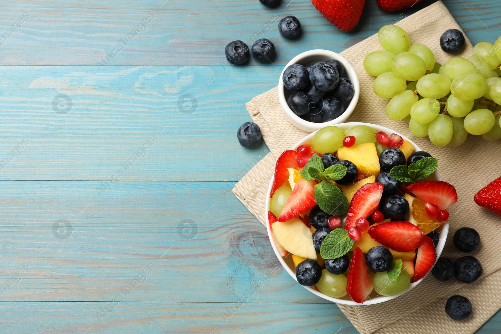 Photo of Tasty fruit salad in bowl and ingredients on light blue wooden table, flat lay. Space for text