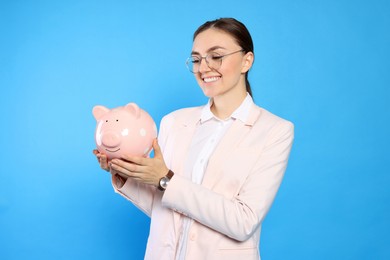 Happy young businesswoman with piggy bank on light blue background