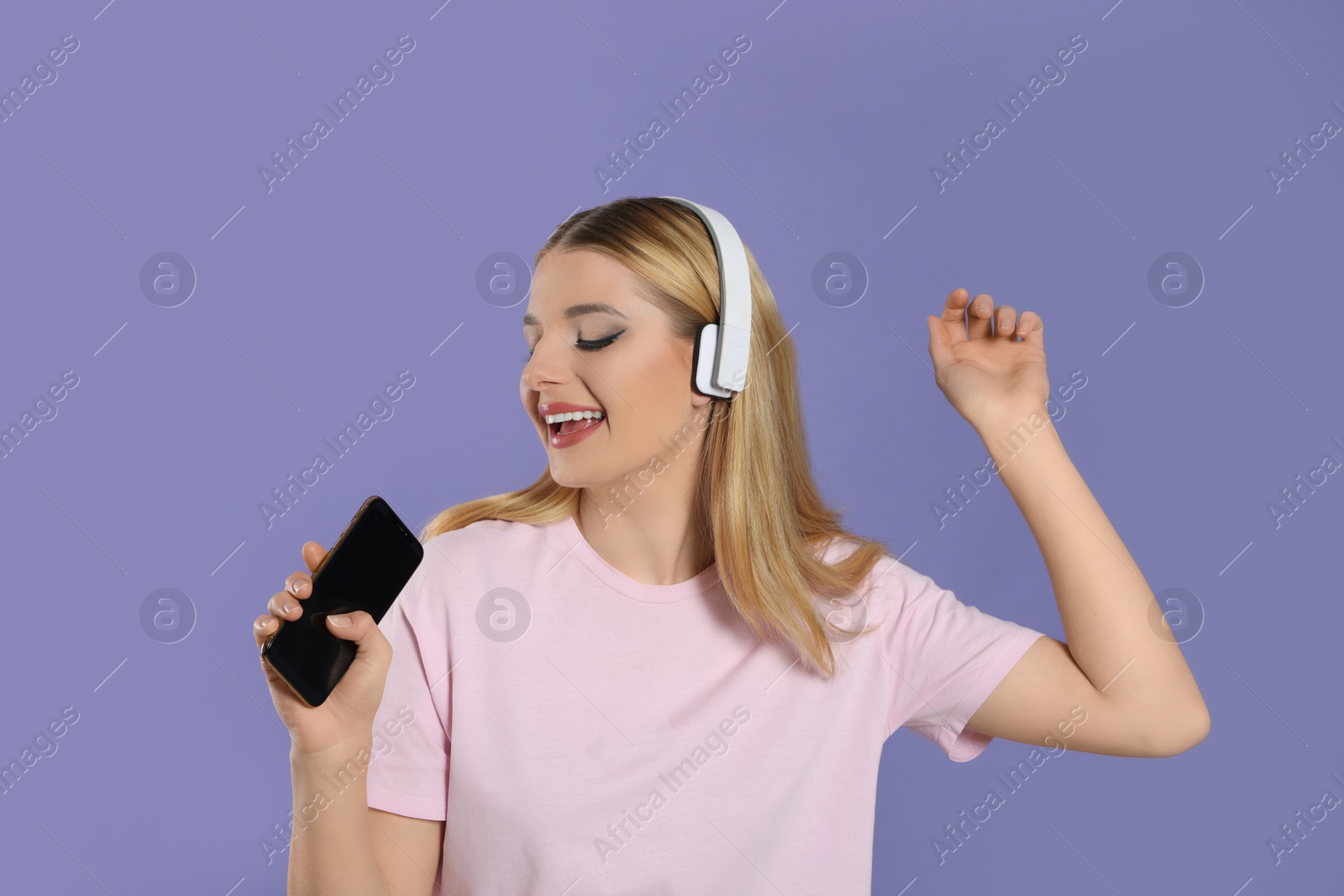 Photo of Happy woman in headphones enjoying music and singing into smartphone on violet background