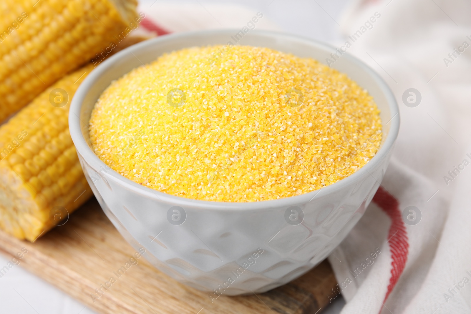 Photo of Raw cornmeal in bowl and corn cobs on white table, closeup