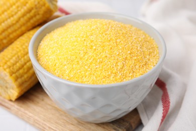 Raw cornmeal in bowl and corn cobs on white table, closeup
