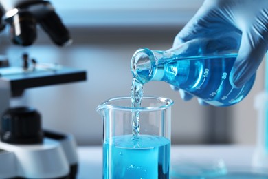 Photo of Scientist pouring light blue liquid from flask into beaker in laboratory, closeup