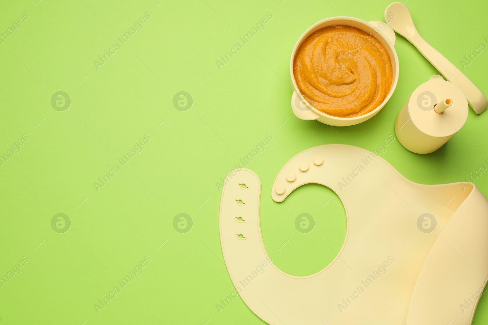 Photo of Silicone baby bib and plastic dishware with healthy food on light green background, flat lay. Space for text