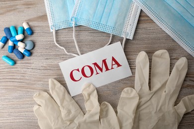 Photo of Card with word Coma, pills, gloves and surgical masks on wooden table, flat lay