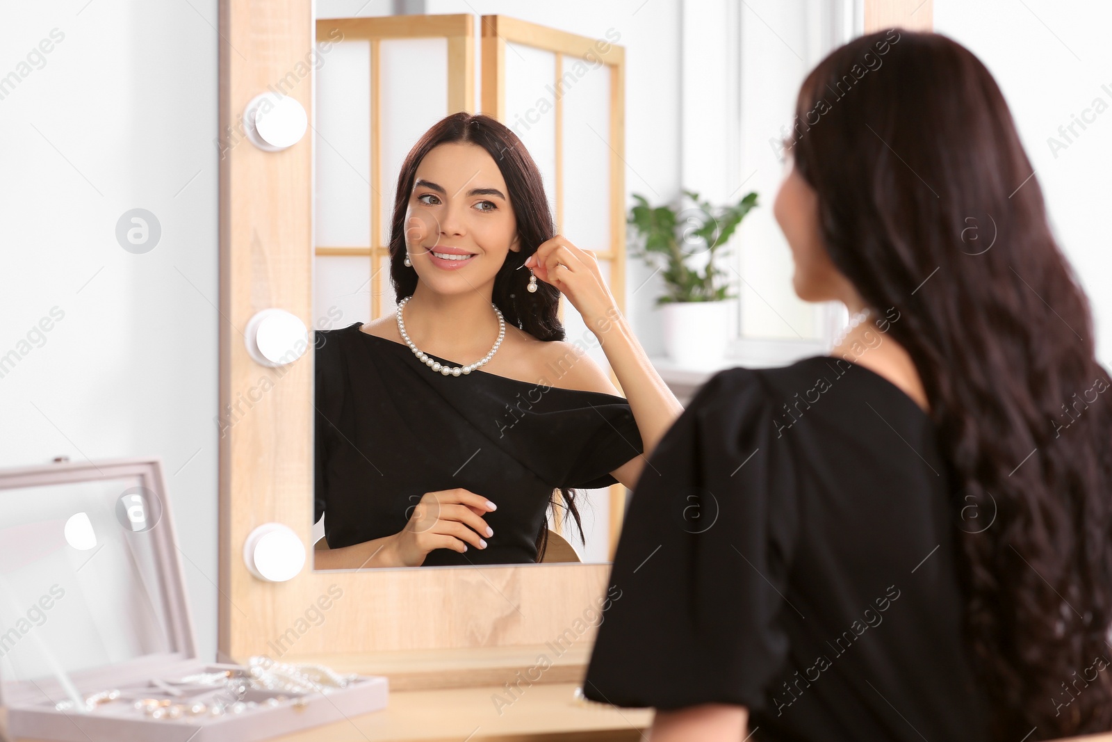Photo of Young woman trying on elegant pearl earring near mirror indoors