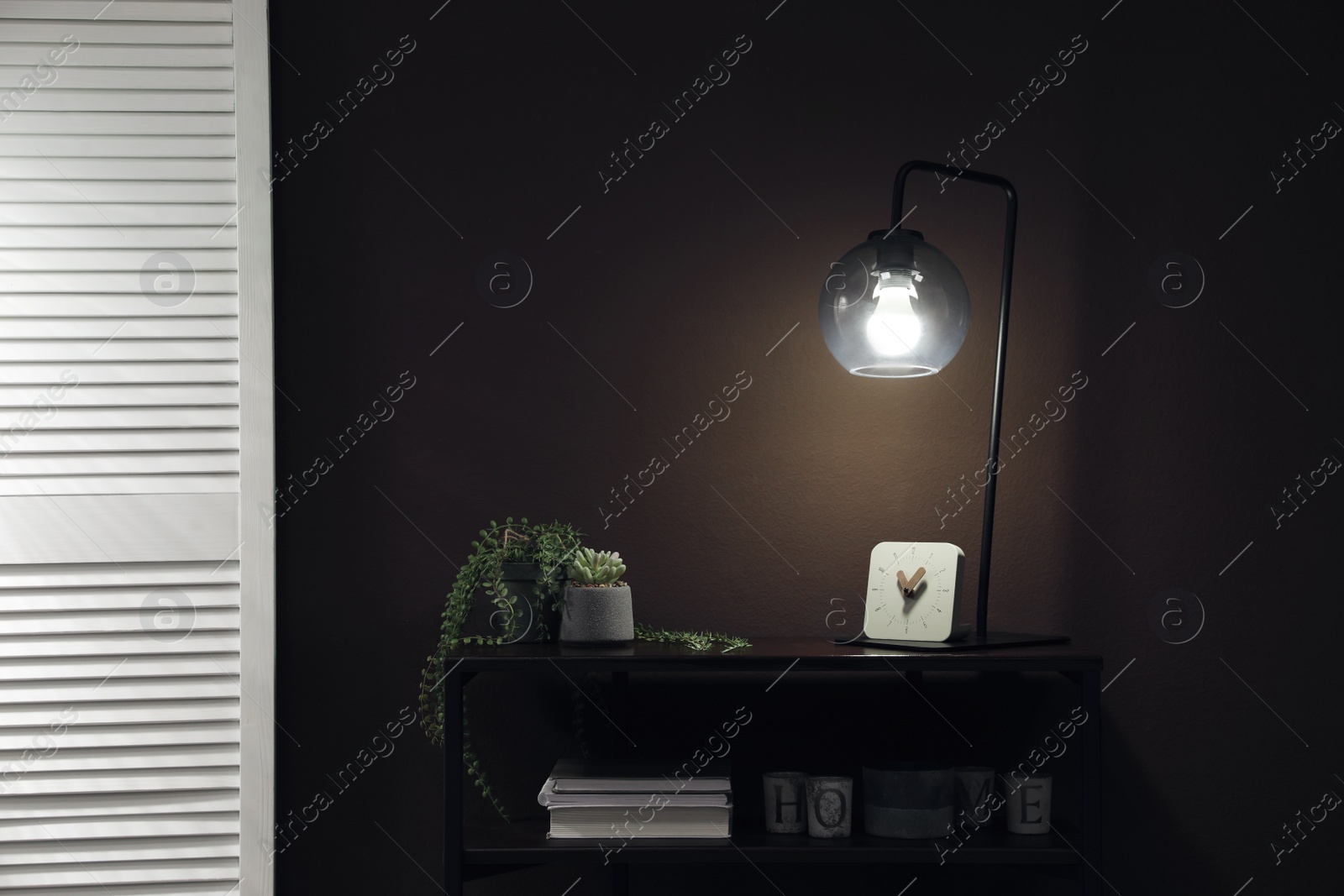 Photo of Stylish lamp, green plants and other stuff on table near brown wall indoors. Interior design