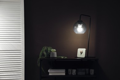 Stylish lamp, green plants and other stuff on table near brown wall indoors. Interior design