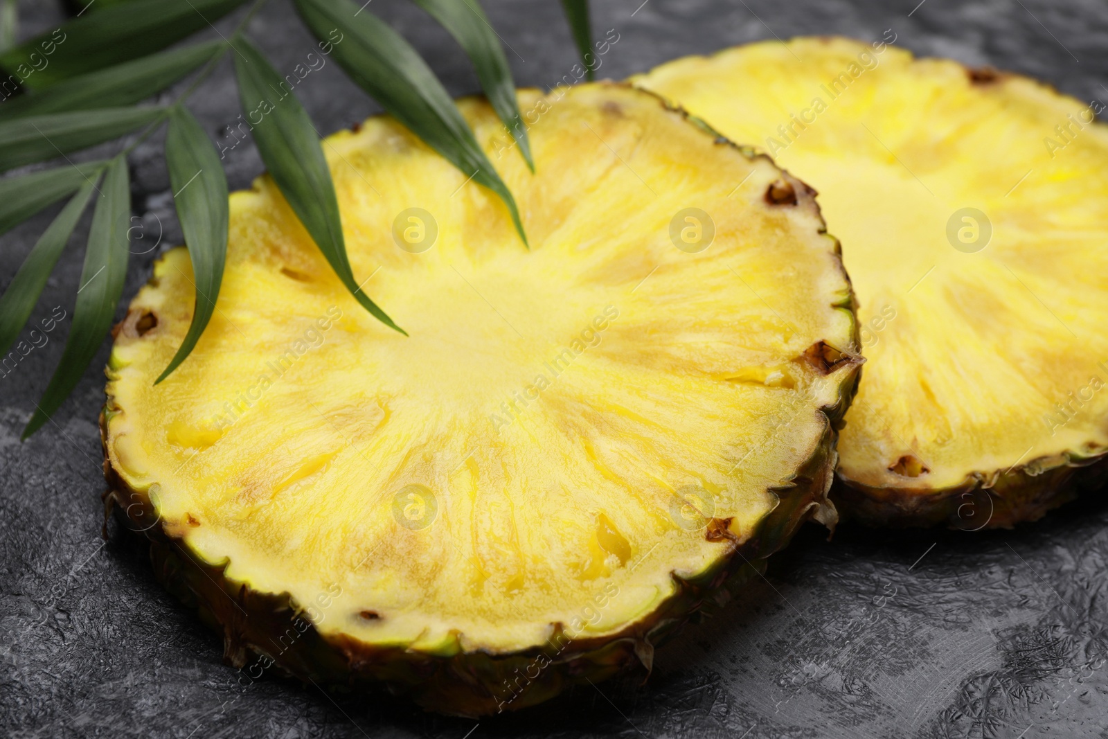 Photo of Slices of tasty ripe pineapple and green leaves on black textured table, closeup