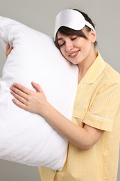 Photo of Happy woman in pyjama and sleep mask holding pillow on grey background