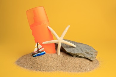 Photo of Sand with bottle of sunscreen, starfish, stone and ship figure against orange background. Sun protection
