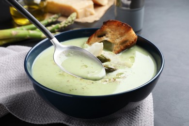 Photo of Spoon with delicious asparagus soup over bowl on dark table