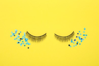 Photo of False eyelashes and shiny confetti on yellow background, flat lay