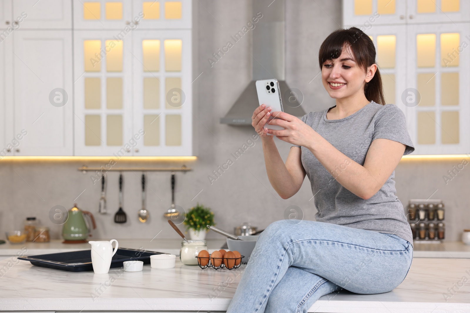 Photo of Happy young housewife using smartphone on white marble table in kitchen