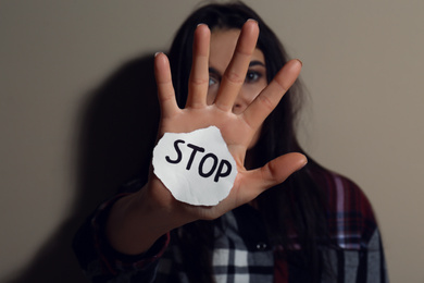 Abused young woman with sign STOP near beige wall, focus on hand. Domestic violence concept