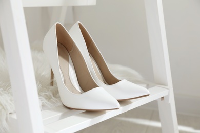 Photo of Pair of white wedding high heel shoes on wooden rack indoors, closeup