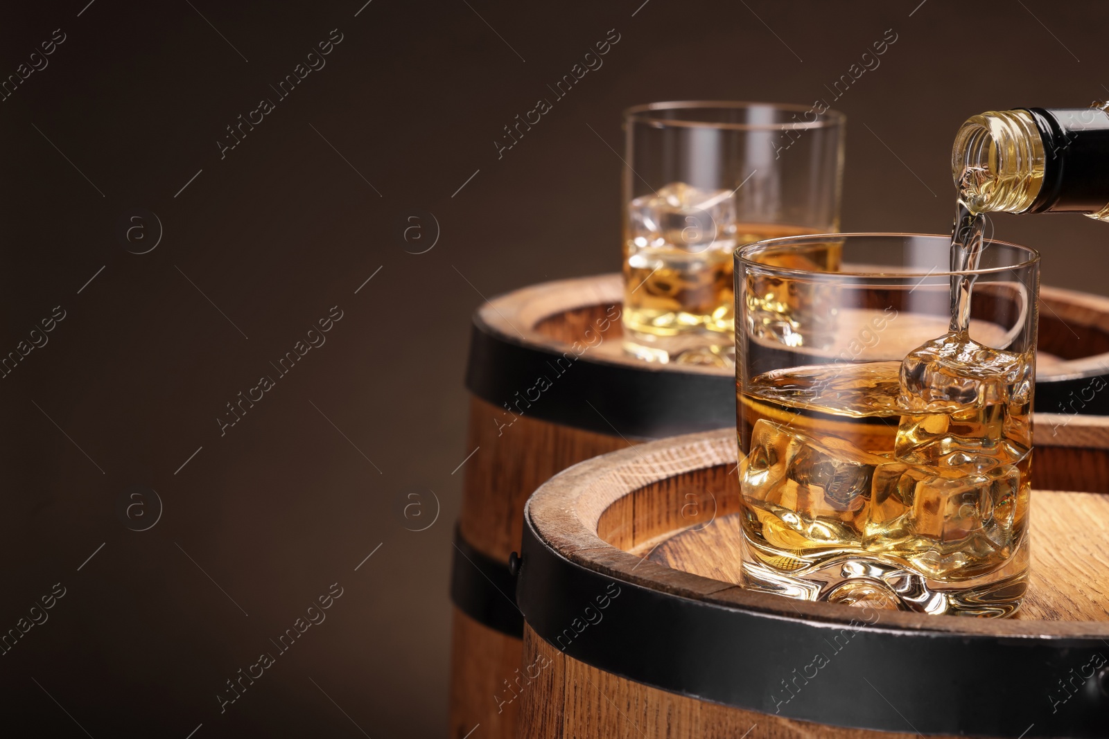 Photo of Pouring whiskey from bottle into glass on wooden barrel against brown background, closeup. Space for text