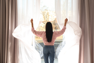 Woman opening window curtains at home in morning, back view