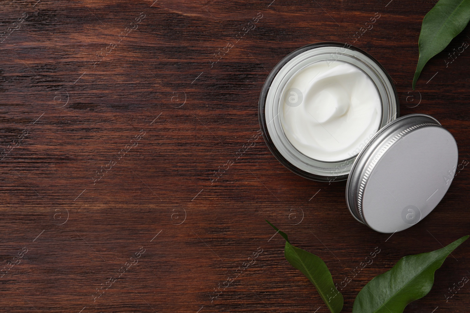 Photo of Jar of face cream and green leaves on wooden table, flat lay. Space for text