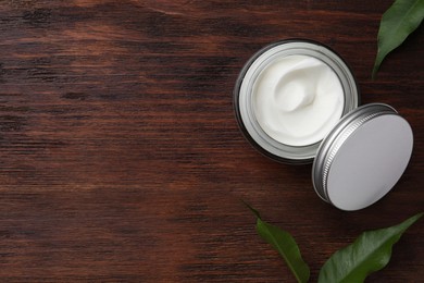 Jar of face cream and green leaves on wooden table, flat lay. Space for text