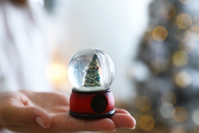 Young woman holding snow globe with Christmas tree on blurred background, closeup