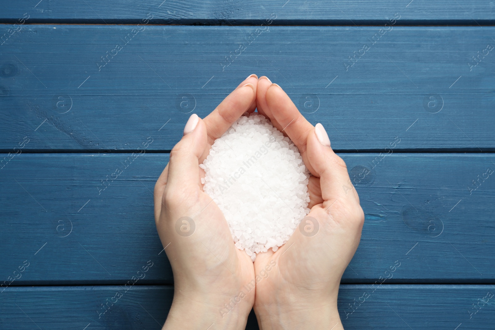 Photo of Woman holding pile of natural sea salt at blue wooden table, top view