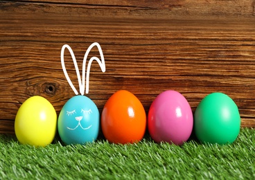 One egg with drawn face and ears as Easter bunny among others on green grass against wooden background