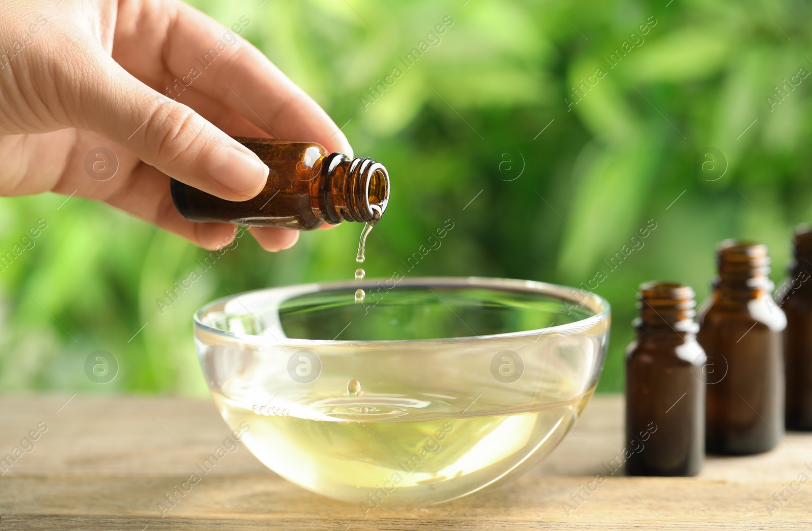 Photo of Woman pouring essential oil from glass bottle into bowl on table, closeup