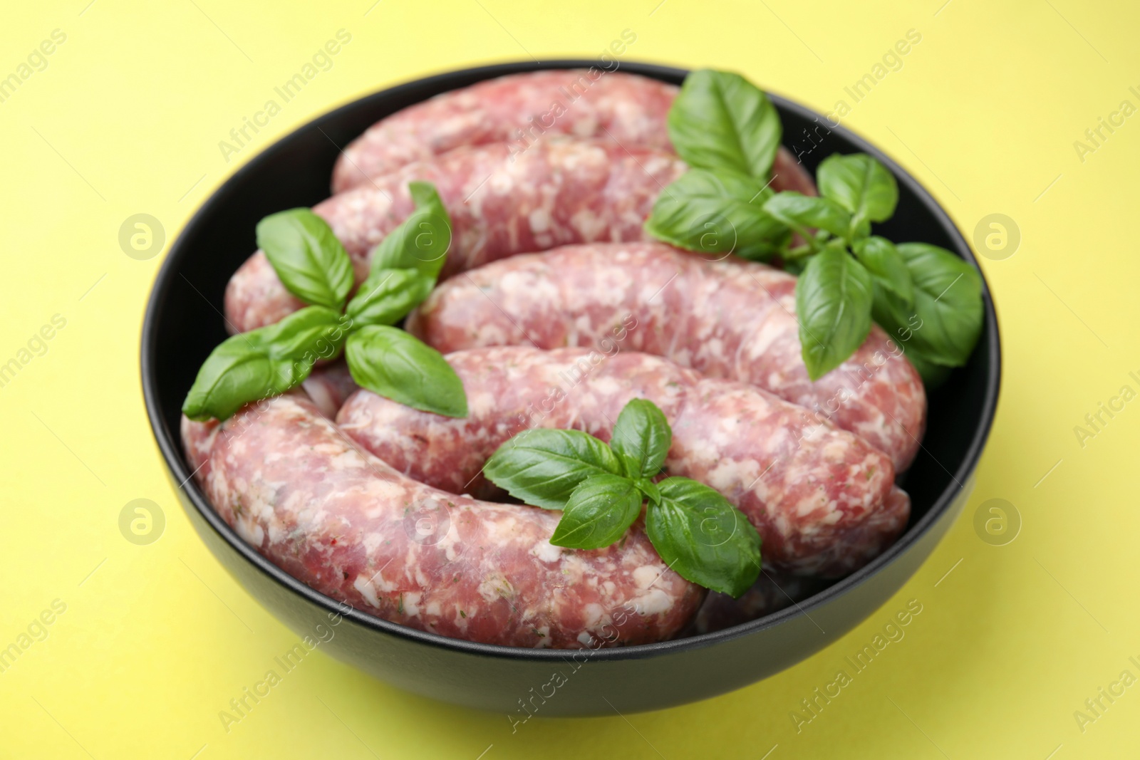 Photo of Raw homemade sausages and basil leaves in bowl on yellow background