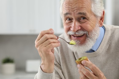 Photo of Happy senior man eating kiwi with spoon indoors. Space for text