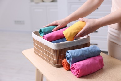 Photo of Woman putting rolled shirt into basket at table in room, closeup. Organizing clothes