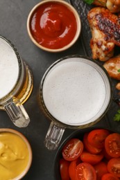 Mugs with beer, delicious baked chicken wings and sauces on dark grey table, flat lay