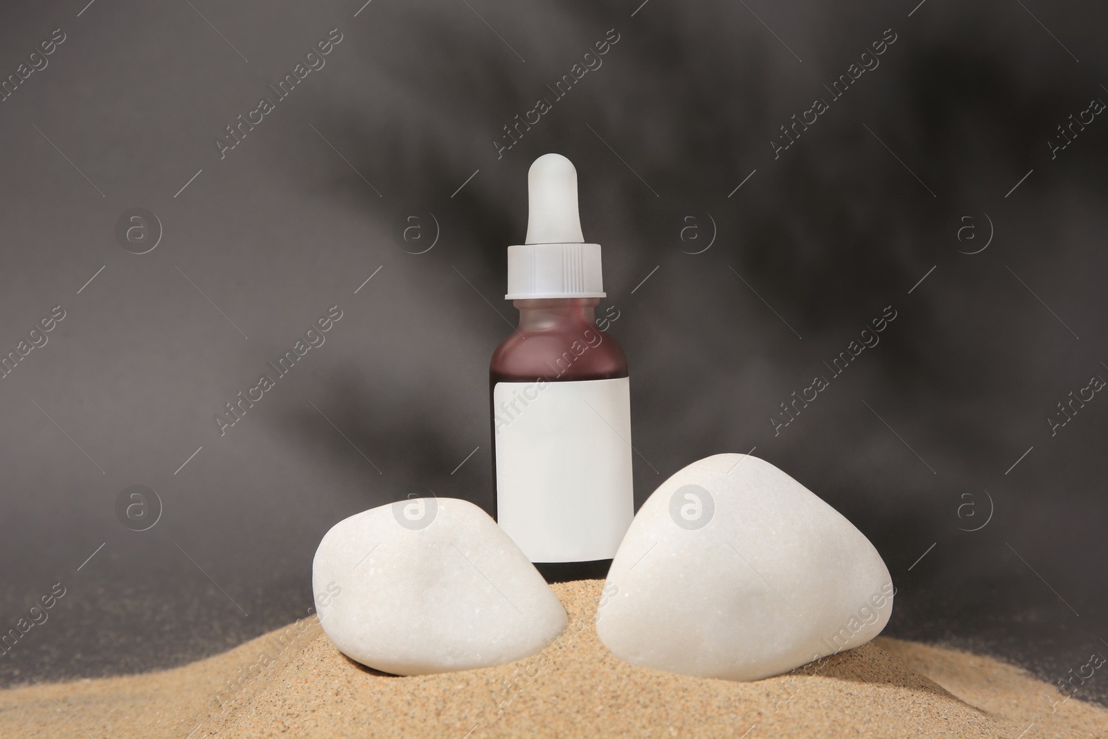 Photo of Bottle of serum and stones on sand against grey background