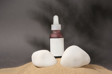 Bottle of serum and stones on sand against grey background