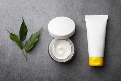 Photo of Jar and tube of face cream with green leaves on grey table, flat lay