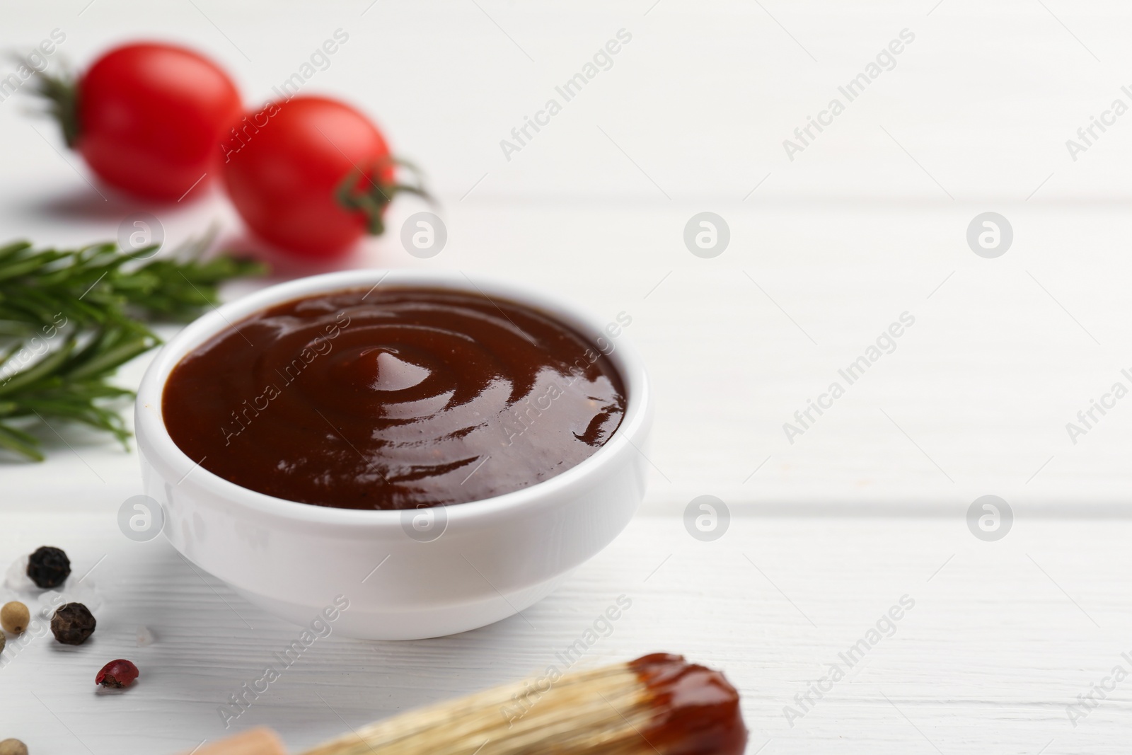 Photo of Tasty barbeque sauce in bowl, rosemary and spices on white wooden table, closeup. Space for text