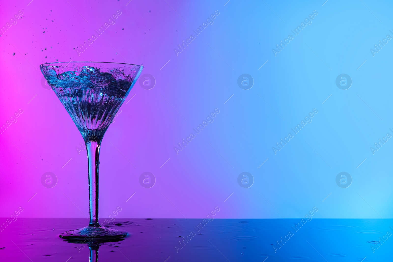 Photo of Glass of martini and splashes on table in neon lights, space for text