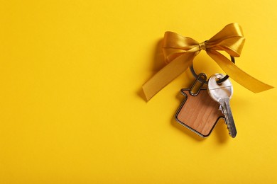 Photo of Key with trinket in shape of house and bow on yellow background, top view. Space for text. Housewarming party