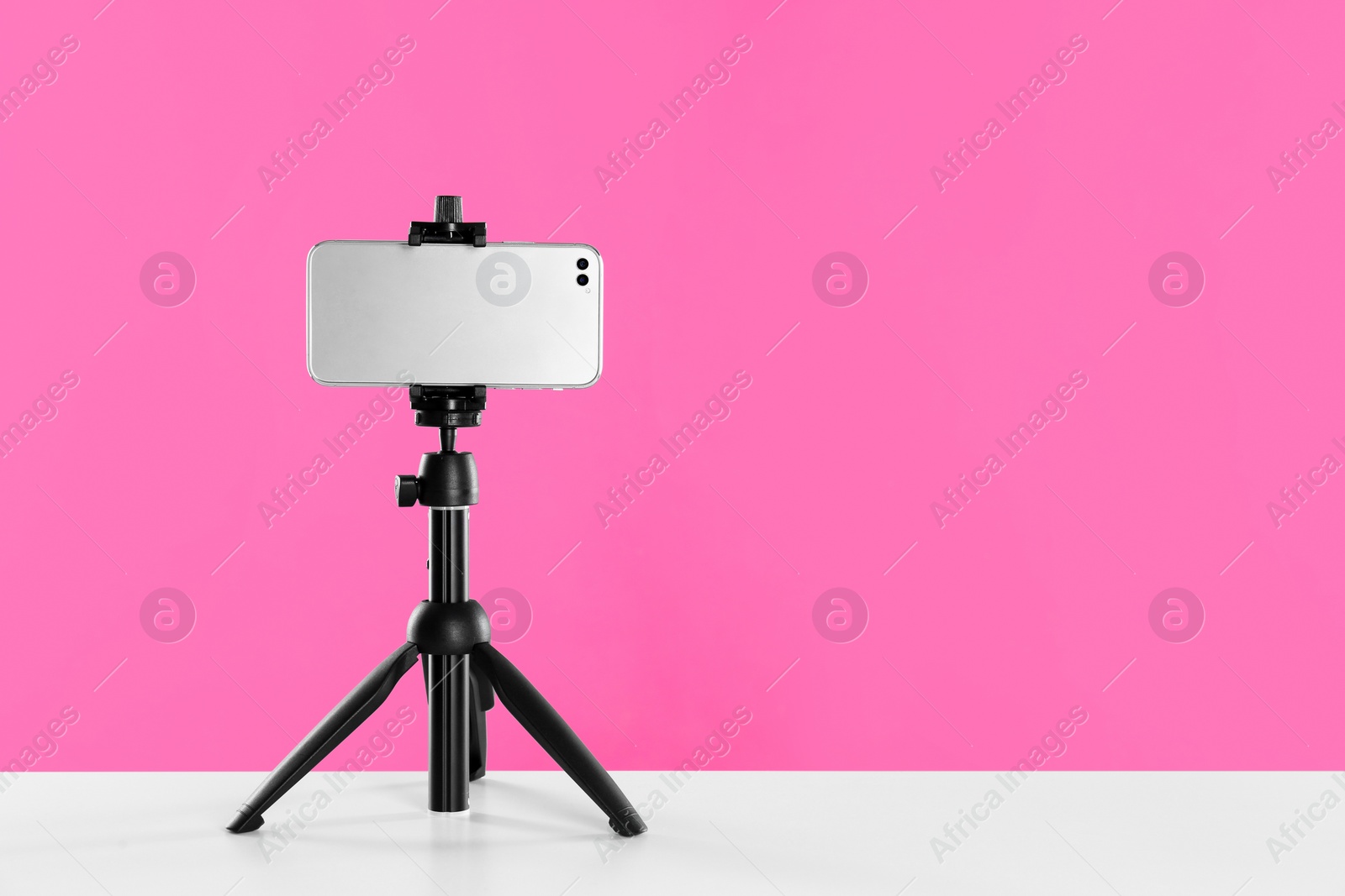 Photo of Smartphone fixed to tripod on white table against pink background. Space for text