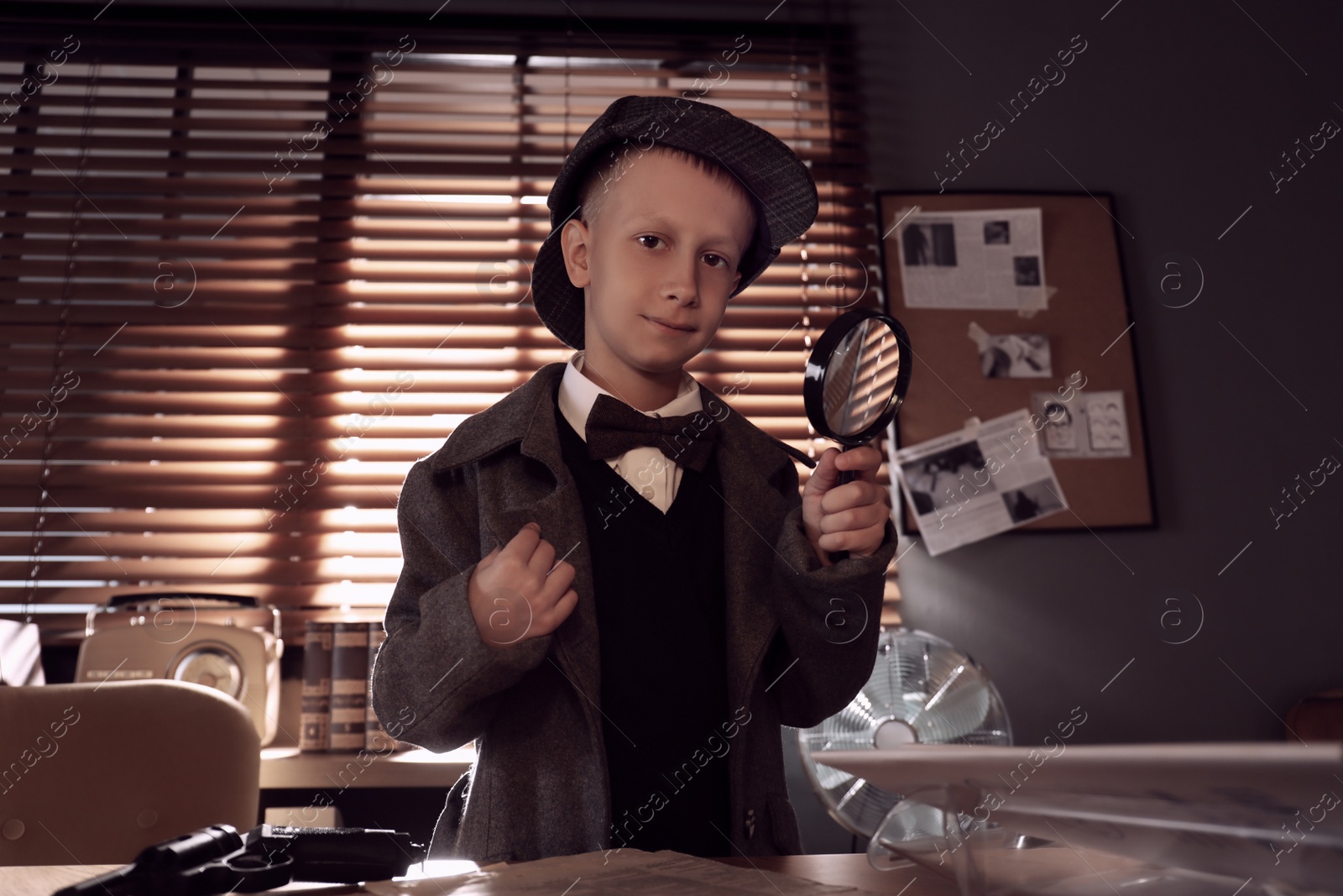 Photo of Cute little detective with magnifying glass at table in office