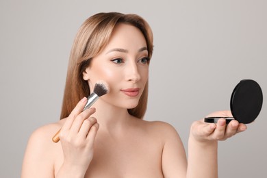 Beautiful young woman applying face powder with brush on grey background