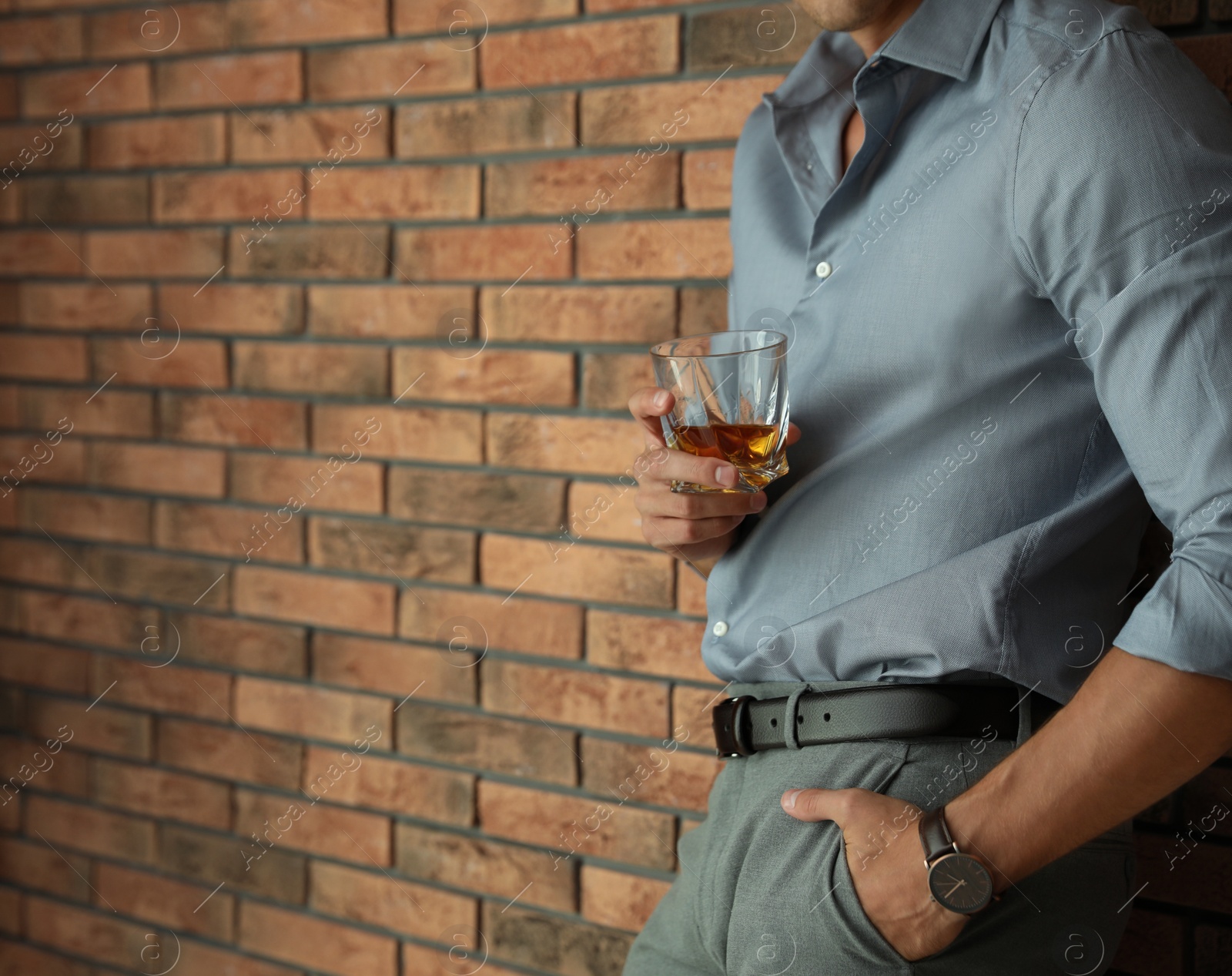 Photo of Young man with glass of whiskey near brick wall indoors, closeup. Space for text