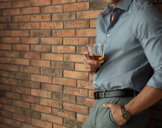 Young man with glass of whiskey near brick wall indoors, closeup. Space for text