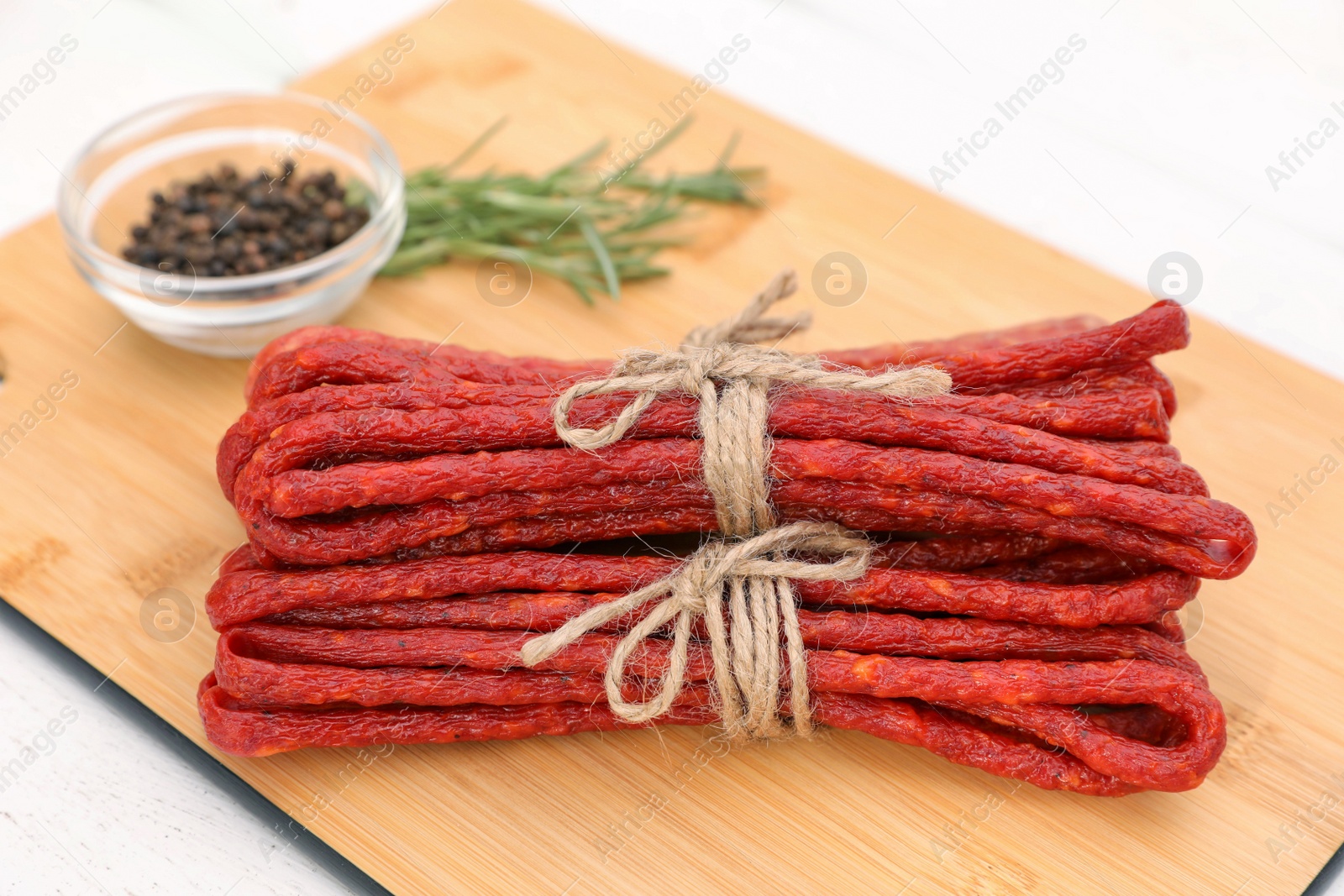 Photo of Bundles of delicious kabanosy with peppercorn and rosemary on white wooden table, closeup